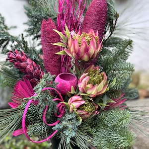Könnte beinhalten: Ein lebendiges Blumenarrangement mit tiefen Magenta- und grünen Elementen. Der Strauß enthält kegelförmige Blüten, artischockenartige Blumen und immergrüne Zweige, akzentuiert mit leuchtend rosa Dekorationselementen. Das Arrangement steht in einer dunkelgrünen Vase.