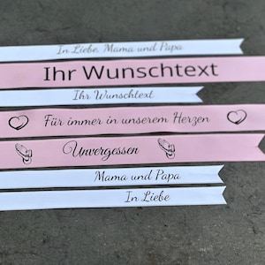 Könnte beinhalten: Rosa und weiße Schleifenbanner mit verschiedenen Botschaften, darunter "In Liebe. Mama und Papa", "Ihr Wunschtext", "Für immer in unserem Herzen", "Unvergessen" und "Mama und Papa".