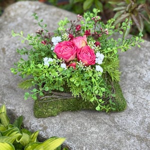 Könnte beinhalten: Eine quadratische, mit Moos bedeckte Basis zeigt ein dekoratives Arrangement aus leuchtend rosa Rosen, kleinen weißen Blüten und dunkelroten Knospen. Die Basis ist mit dünnen braunen Zweigen umwickelt und von dichtem, grünem Blattwerk umgeben.