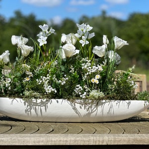 Op de afbeelding: Een witte keramische trog gevuld met verschillende witte bloemen en groen. De trog staat op een houten tafel.