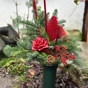 May include: A dark green vase displays a festive arrangement of evergreen branches, red berries, a red rose, and a pine cone. Tall red accents and a textured red flower add height to this seasonal display.