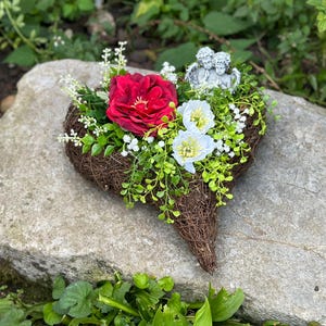 Könnte beinhalten: Ein herzförmiger Kranz aus Zweigen und Ästen, verziert mit einer roten Rose, weißen Blumen und zwei kleinen Engelsfiguren. Der Kranz liegt auf einem grauen Stein.