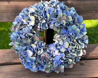 May include: A circular wreath constructed from numerous hydrangea blossoms in varying shades of blue, ranging from deep navy to light sky blue, with subtle hints of muted green and purple tones throughout the dense, layered floral arrangement.