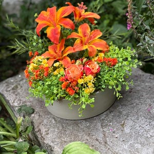 May include: A floral arrangement in a grey ceramic bowl. The arrangement features vibrant orange lilies, orange and yellow roses, and green foliage. The bowl sits on a grey stone surface, with greenery in the background.