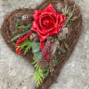Könnte beinhalten: Ein herzförmiger Kranz aus geflochtenen braunen Zweigen, verziert mit einer großen roten Rose, Tannenzapfen und Zweigen mit Grün und roten Beeren. Eine rustikale, natürliche Dekoration.