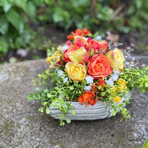 Può includere: Un arrangiamento floreale in un vaso di cemento grigio. L'arrangiamento comprende rose gialle, arancioni e rosse con fogliame verde.