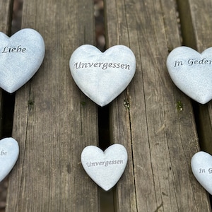 Może przedstawiać: Sześć szarych kamieni w kształcie serca z wygrawerowanymi słowami "In Liebe", "Unvergessen", "In Gedenken". Kamienie są ułożone w dwóch rzędach po trzy na drewnianej powierzchni.