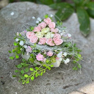 Könnte beinhalten: Ein kleiner, weißer Keramikbehälter, der mit einem zarten Arrangement aus Miniaturrosen in zarten Rosa- und Cremetönen überquillt. Die Rosen sind zwischen Zweigen von Grün und weißen, fadenartigen Akzenten eingebettet und schaffen eine bezaubernde Blumendekoration.