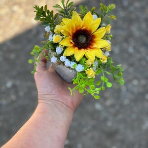 May include: A miniature floral arrangement with a large yellow sunflower and a dark brown centre. The arrangement includes small white and yellow flowers, and green foliage, set in a small, grey, textured pot.