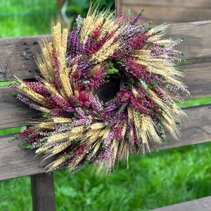 May include: A circular wreath constructed from dried golden wheat stalks interspersed with vibrant purple, magenta, and white heather flowers. The arrangement features a dense, textured composition of natural botanical elements arranged in a radial pattern.