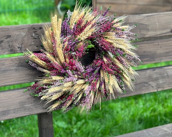 Könnte beinhalten: Ein kreisförmiger Kranz aus getrockneten goldenen Weizenhalmen, durchsetzt mit leuchtend violetten, magentafarbenen und weißen Heidekrautblüten. Das Arrangement zeigt eine dichte, strukturierte Komposition aus natürlichen botanischen Elementen.
