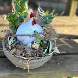 Può includere: Una statuetta di pollo in ceramica bianca con cresta e bargigli rossi è posizionata in una ciotola grigia piena di ramoscelli marroni, foglie verdi e piume. Un uovo marrone macchiato è annidato tra i ramoscelli.