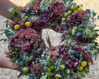 Op de afbeelding: Een ronde bloemenkrans samengesteld uit donkerpaarse hortensiabloemen, felroze heide, gele craspedia-bollen, blauwe distels, groene bessen en zilverkleurig blad. Het arrangement bevat een dichte mix van verschillende texturen en kleuren.
