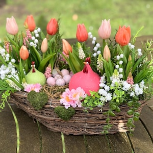 May include: An Easter basket arrangement with tulips, white flowers, and greenery. The basket contains two ceramic chickens and small eggs. The basket is made of woven branches. The tulips are in shades of pink and orange.