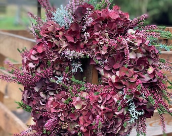 Op de afbeelding: Een ronde bloemenkrans met diep bordeauxrode hortensiabloesems, afgewisseld met felroze heidetakjes en zilvergroen blad. Het arrangement is dicht op elkaar geplaatst, wat zorgt voor een rijke, getextureerde weergave van botanische elementen in herfsttinten.