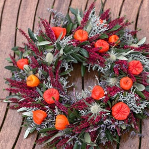 Pode incluir: Uma coroa circular com lanternas de physalis em laranja vibrante, ramos de urze em magenta profundo, folhagem verde prateada e cabeças de cardo espinhosas. O arranjo é densamente preenchido com texturas e cores variadas.