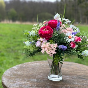 Peut inclure: Un bouquet de fleurs roses, blanches et violettes dans un vase en verre transparent. Les fleurs sont disposées dans un vase sur une table en bois. L'arrière-plan est un champ vert.