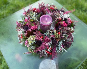 May include: A circular floral wreath featuring deep purple and magenta heather, dark foliage, and bright red berries. A clear glass cylinder sits in the center, holding a solid purple pillar candle with a visible white wick at the top.