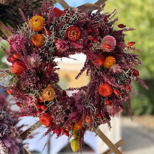 May include: A circular wreath composed of dried purple heather, vibrant orange strawflowers, and small red berries. The arrangement features a dense texture with interspersed dried foliage and botanical elements, creating a rich, multi-colored display.