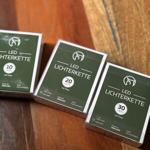 May include: Three boxes of LED string lights with the text "LED LICHTERKETTE" in white on a green background. The boxes display 10, 20, and 30 LEDs, respectively. Each box includes the text "mit Timer" and other details.
