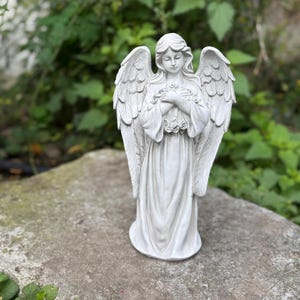 Könnte beinhalten: Eine weiße Engelsstatue mit detaillierten Flügeln und einem ruhigen Ausdruck, die Blumen hält. Die Statue steht auf einer Steinoberfläche mit einem Hintergrund aus grünem Laub.
