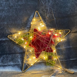 Könnte beinhalten: Goldfarbene Draht-Stern-Weihnachtsdekoration mit warmweißen LED-Lichtern. Der Stern hat eine rote Samtmitte und ist mit Tannennadeln und kleinen Tannenzapfen verziert. Auf grauem Hintergrund.