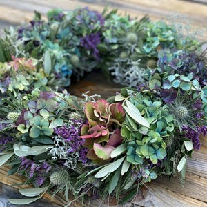 Pode incluir: Uma grinalda floral circular composta por diversas flores e folhagens secas. A grinalda apresenta hortênsias em tons de verde, azul e roxo, juntamente com raminhos de lavanda, cardo e folhas de oliveira. A grinalda é exibida sobre uma superfície de madeira.