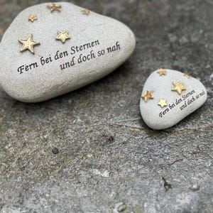 Könnte beinhalten: Zwei dekorative, hellgraue Steine mit goldenen Sternverzierungen. Der größere Stein trägt den Text "Fern bei den Sternen und doch so nah". Der kleinere Stein hat die gleiche Inschrift. Die Steine liegen auf einer grauen Oberfläche.