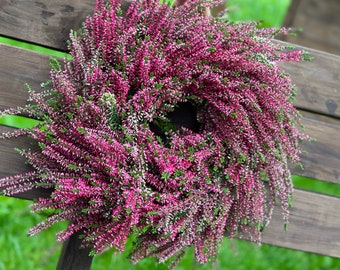 Könnte beinhalten: Ein kreisförmiger Kranz aus dichten Büscheln leuchtend magentafarbener und dunkelrosa Heidekrautblüten mit kleinen grünen Blättern. Das Blumenarrangement ist in einer vollen, strukturierten Ringform angeordnet und zeigt die natürlichen Stängel.