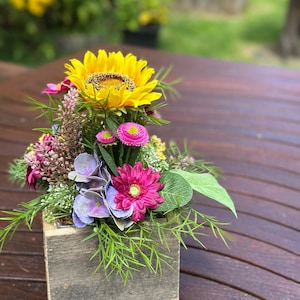 Könnte beinhalten: Ein Blumenarrangement in einer quadratischen, verwitterten Holzkiste mit einer großen gelben Sonnenblume, leuchtend pinken Gänseblümchen, violetten Hortensienblüten und verschiedenen grünen Blattzweigen. Das Gesteck besteht aus mehreren Blumenarten.