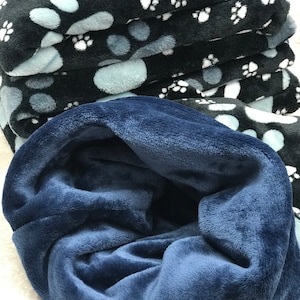 Könnte beinhalten: Ein Stapel schwarzer und blauer Fleece-Decken mit einem weißen Pfotenabdruckmuster. Die oberste Decke ist gefaltet, um ein einfarbiges blaues Fleece-Innenfutter zu zeigen.