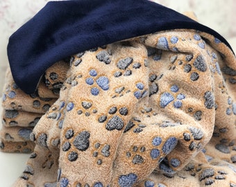 Könnte beinhalten: Eine weiche, flauschige Decke mit einem Pfotenabdruckmuster in Beige, Grau und Blau und einem marineblauen Rand. Die Decke ist gefaltet und zeigt das Pfotenmuster und den marineblauen Stoff.