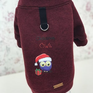 Könnte beinhalten: Burgunderroter Hundepullover mit einem Weihnachts-Eulen-Design. Die Eule trägt einen Weihnachtsmann-Hut und hält ein Geschenk. Der Pullover hat einen schwarzen Riemen mit einem Metallring.