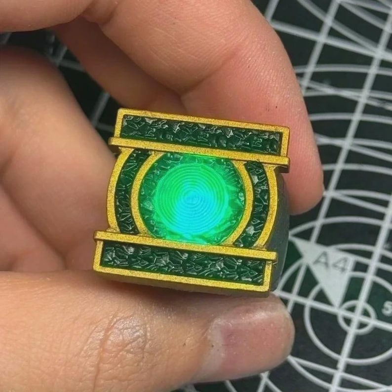 May include: A ring featuring a rectangular gold-colored frame with textured dark green panels and a central circular glowing green light. The design includes a prominent circular emblem with concentric rings, set against a dark green background.