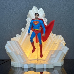 Peut inclure: Une figurine Superman dans un costume bleu et rouge classique avec une cape rouge, posée devant un présentoir lumineux blanc et géométrique. Le présentoir a un design cristallin, et la figurine est suspendue en l'air.