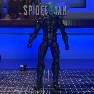 May include: A blue-skinned action figure wearing a black suit with a silver chest piece. The figure is standing on a black and white grid surface.