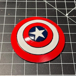 Pode incluir: Um escudo do Capitão América vermelho, branco e azul com uma estrela de cinco pontas no centro. O escudo é feito de plástico e tem um acabamento brilhante.