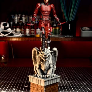Pode incluir: Uma figura de ação vermelha de um super-herói com um fato e máscara vermelhos, segurando bastões vermelhos, em pé sobre uma estátua de gárgula no topo de uma estrutura de tijolos. A estátua é cinzenta e o tijolo é castanho.