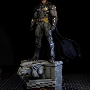 May include: A detailed Batman action figure standing on a stone-textured base featuring a grey gargoyle sculpture. The figure wears a dark grey armoured suit with a gold utility belt and a flowing black cape, positioned against a solid black background.
