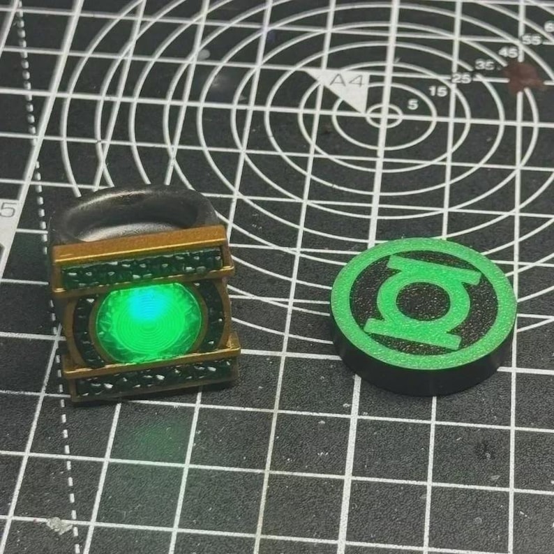 May include: A metallic ring featuring a rectangular gold-toned frame with dark green textured accents and a glowing circular green center, positioned next to a circular black and green disc displaying the Green Lantern symbol on a grid mat.