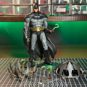 May include: A dark grey and black Batman action figure with a green question mark on his chest. The figure is holding a green question mark in his right hand. The figure is standing on a green base with a question mark on it. The figure is surrounded by several other accessories, including a green question mark, a silver and black trap, and a silver and black dome.