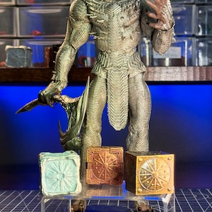 Puede incluir: Una figura de acción plateada y dorada de una criatura con un casco con cuernos y una hoja afilada. La figura está de pie sobre un soporte de exhibición de acrílico transparente con tres cajas pequeñas, una azul, una roja y una dorada.