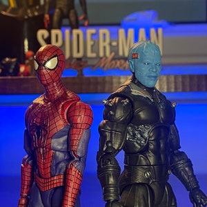May include: Two action figures, one red and blue Spider-Man and one blue and black Doc Ock, stand on a blue surface. The figures are posed in a dynamic action stance. The background features a sign that reads "Spider-Man: No Way Home".
