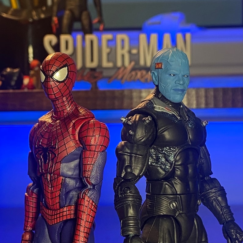 May include: Two action figures, one red and blue Spider-Man and one blue and black Doc Ock, stand on a blue surface. The figures are posed in a dynamic action stance. The background features a sign that reads "Spider-Man: No Way Home".