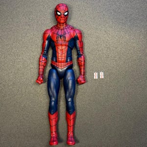 Puede incluir: Una figura de acción de Spider-Man con un traje rojo y azul con un patrón de telaraña negro, un emblema de araña negro en el pecho y lentes oculares blancos. La figura está de pie con dos pequeños accesorios rectangulares blancos a la derecha.