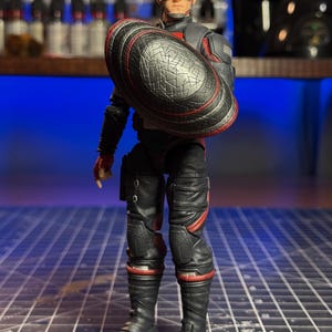 May include: A Captain America action figure in a black and red suit, holding a shield. The figure wears a helmet with a white star and goggles. The shield is black with a red border. The figure is standing on a grid.