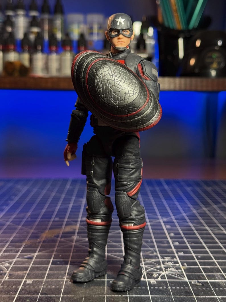 May include: A Captain America action figure in a black and red suit, holding a shield. The figure wears a helmet with a white star and goggles. The shield is black with a red border. The figure is standing on a grid.