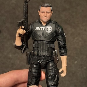 May include: Action figure of a man in a black tactical uniform, holding a black rifle. The figure has the letters "AVTF" and a skull emblem on the chest. The figure is wearing black pants and a tactical vest.