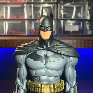 May include: A black and gray action figure of Batman with a black cape and a gold belt. The figure has a detailed face with blue eyes.