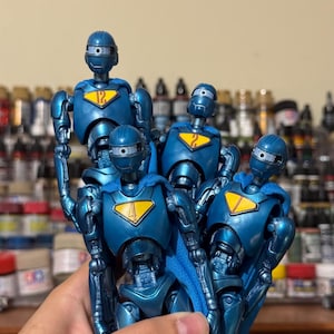 Peut inclure: Cinq figurines de robots bleus métalliques. Chaque figurine porte un numéro unique sur sa poitrine et une cape bleue. Les figurines sont présentées avec les bras levés, mettant en valeur leur design détaillé et leur articulation. Les figurines mesurent environ 15 cm de haut.